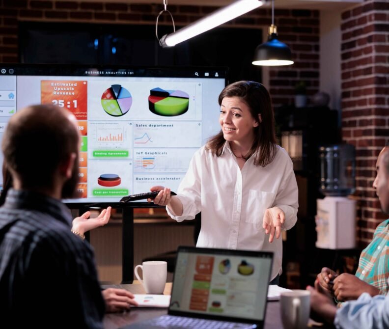 smiling-caucasian-woman-startup-office-doing-business-presentation-big-tv-screen-with-charts-front-team-confident-employee-presenting-growing-sales-statistics-late-night-meeting_11zon